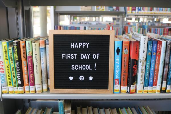 Photo of letterboard with "Happy First Day of School!" sitting on library book shelf. 