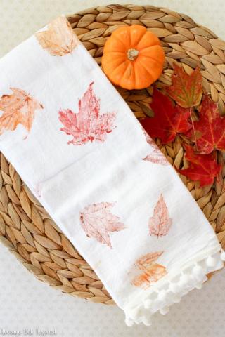 leaf painted tea towel on a wicker stand with a pumpkin and fall leaves beside it.