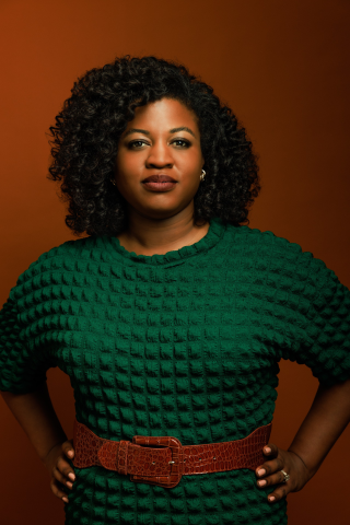 A photo of Dejear with a green dress, brown belt, and orange/red background.